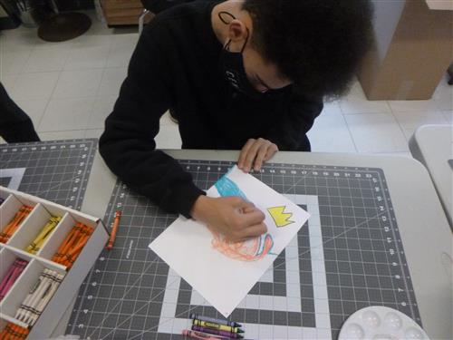 Photo shows a student bent over an art table, using crayons to create artwork in the style of Basquiat. 